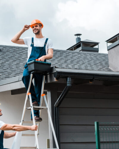 happy repairman sitting on roof and holding toolbox near coworker in helmet